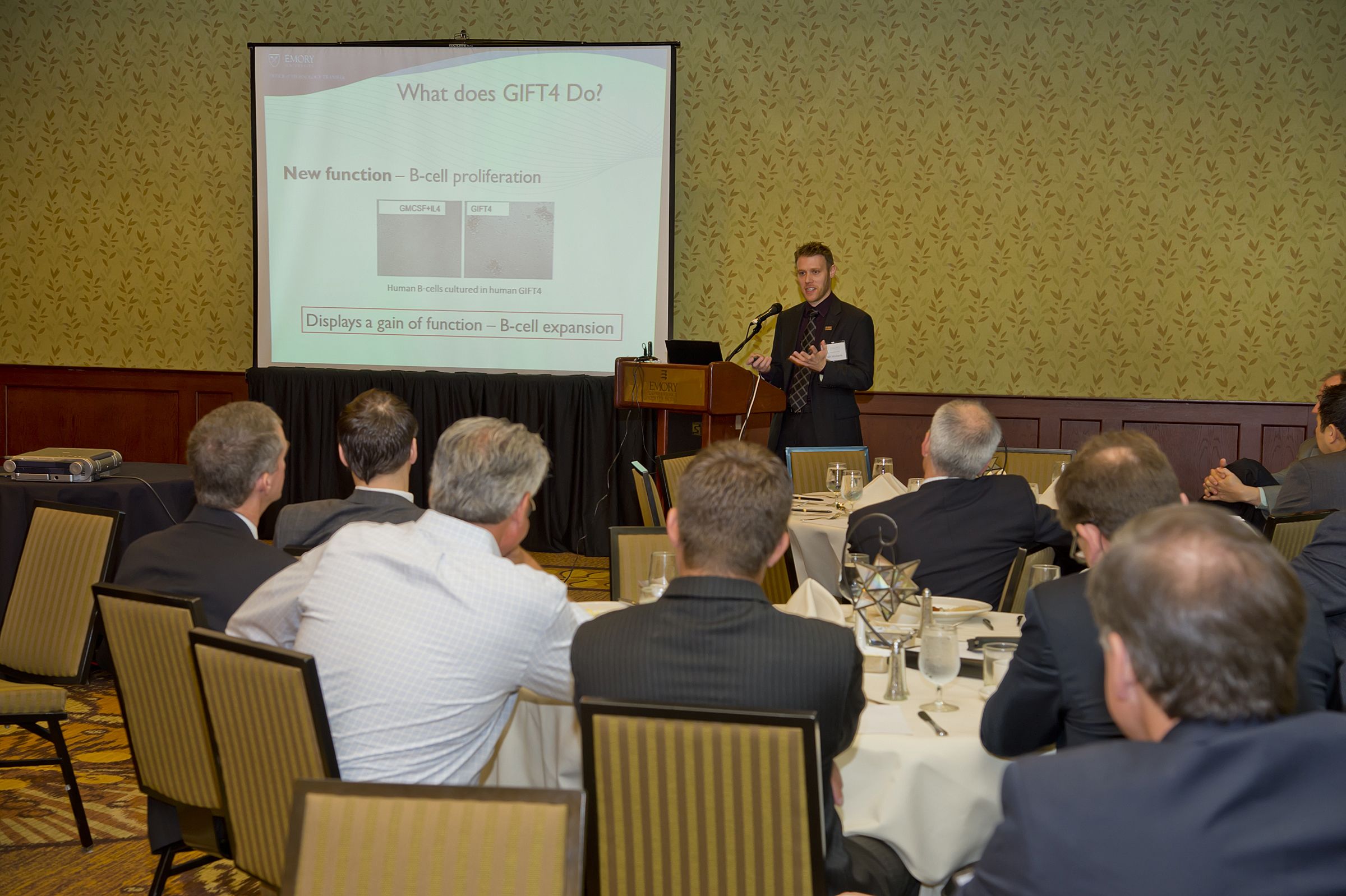 Photo of a speaker at a podium with a screen behind him. People at white-tableclothed tables listen.