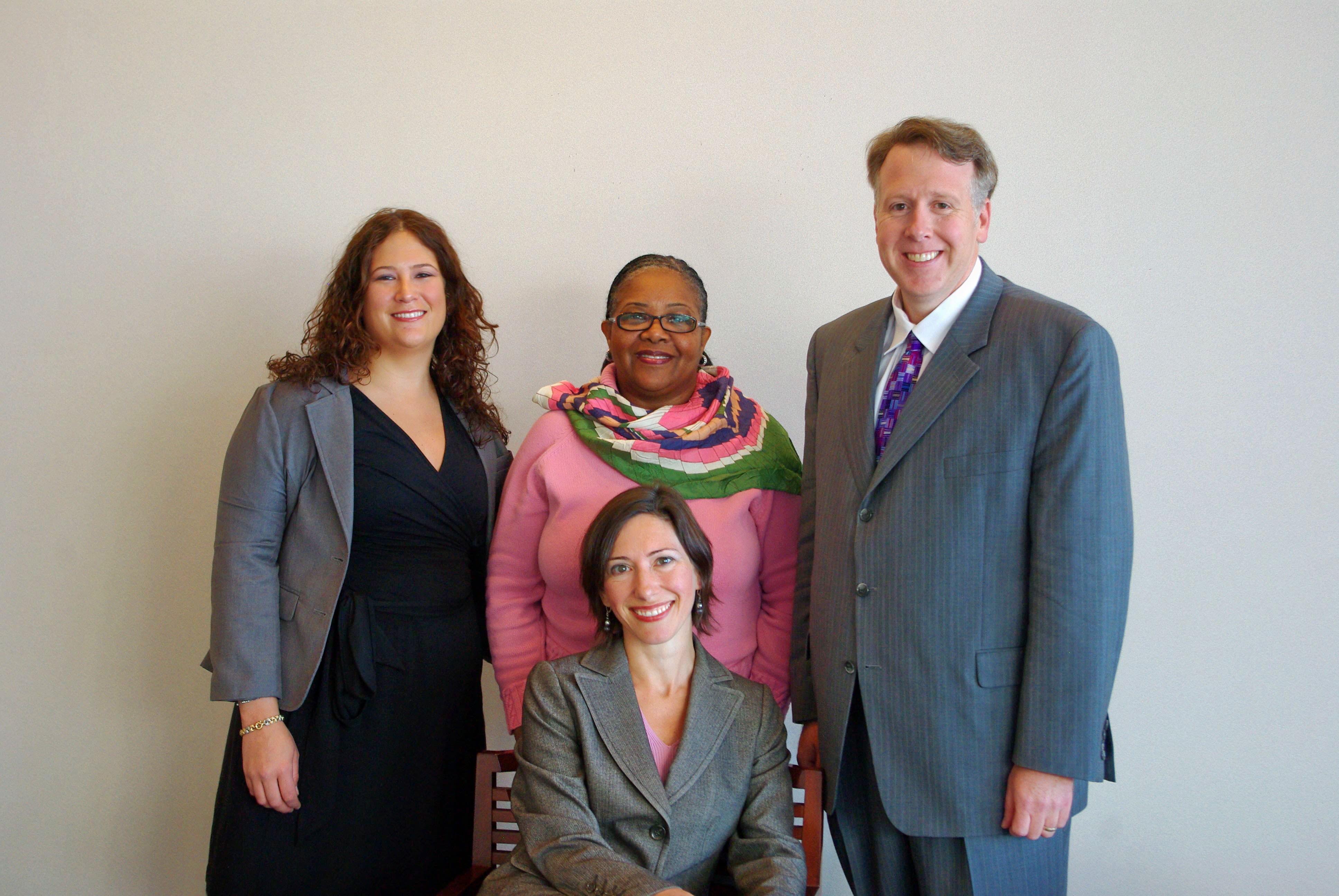 Three people -- two women and one man -- stand behind a seated smiling woman.