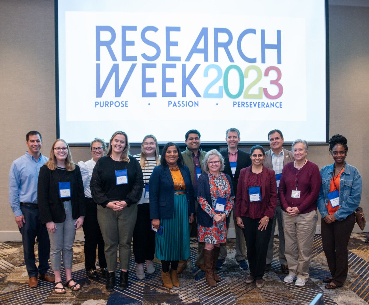 Group of people standing in front of a large screen displaying “Research Week 2023: Purpose, Passion, Perseverance.”