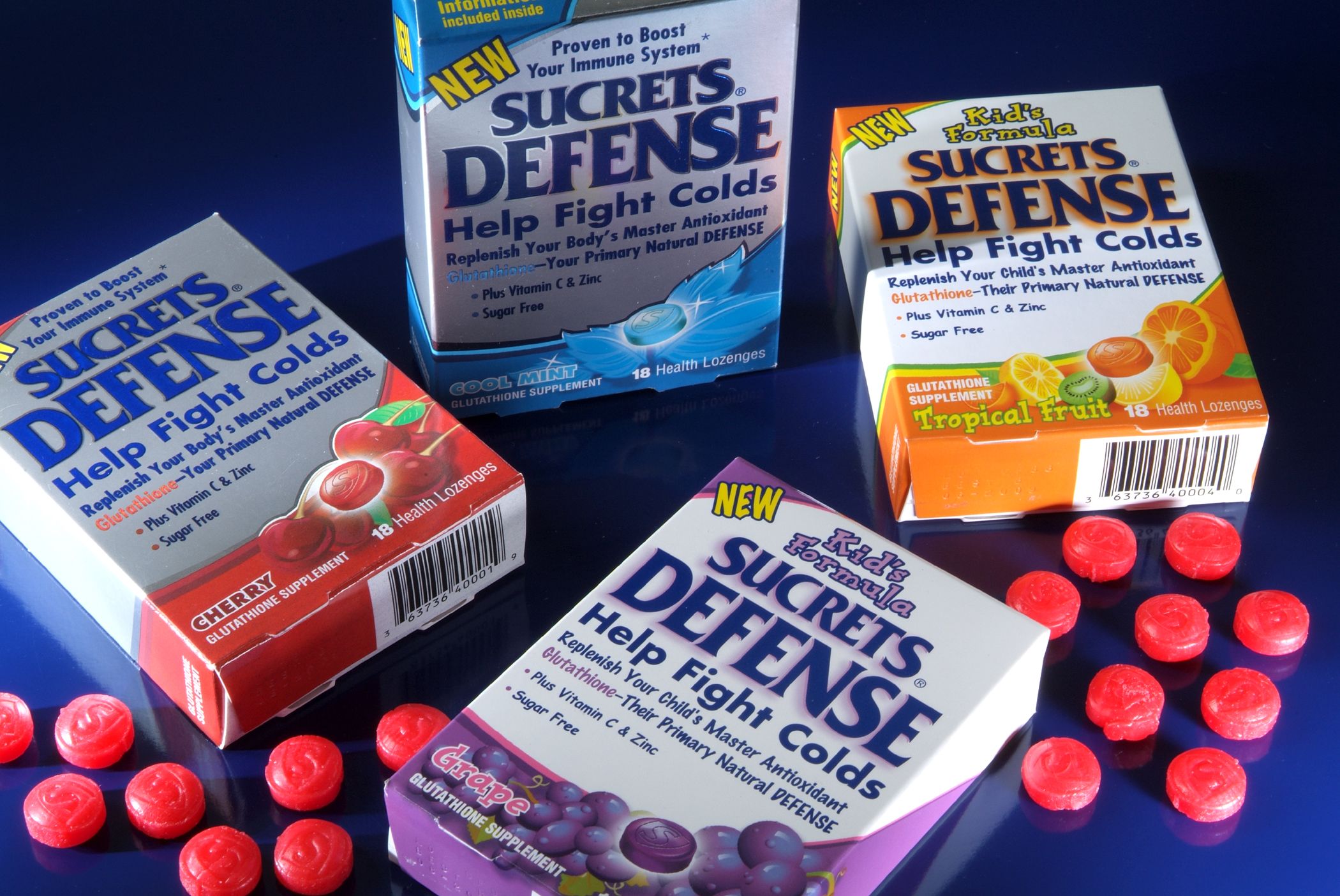 Four Sucrets Defense lozenge boxes in assorted flavors with red lozenges scattered around.
